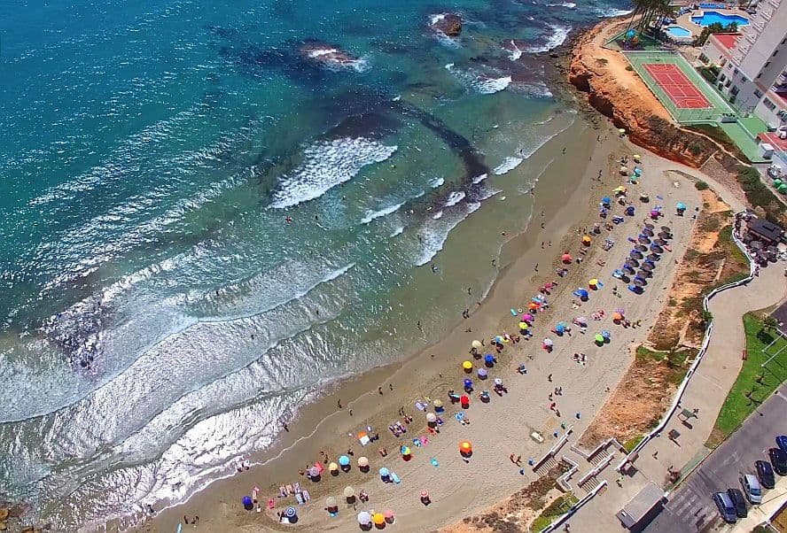 Vista panoràmica de la platja Flamenca amb para-sol, pista de tennis i piscines al costat d'un hotel a la cantonada superior dreta de la imatge.
