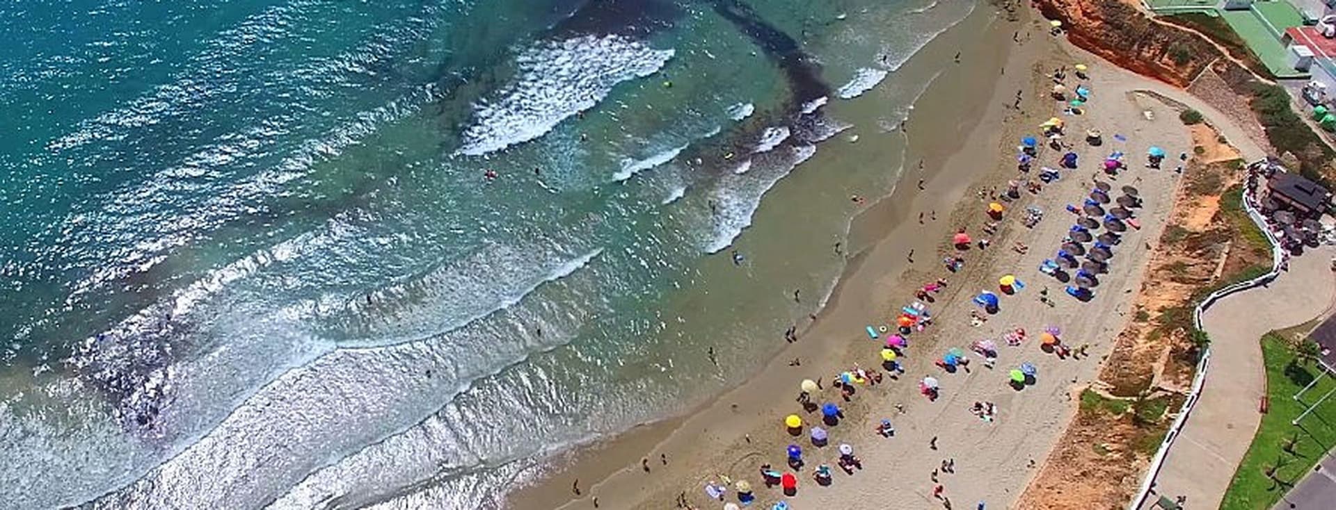 Vista panoràmica de la platja de Platja Flamenca amb para-sol, pista de tennis i piscines al costat d'un hotel a l'angle superior dret de la imatge.