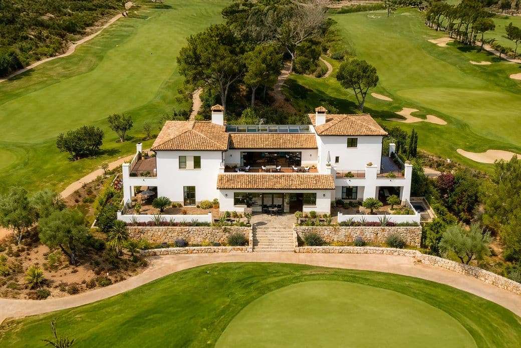 Aerial view of a white villa surrounded by a lush golf course with sand bunkers and trees on a sunny day.