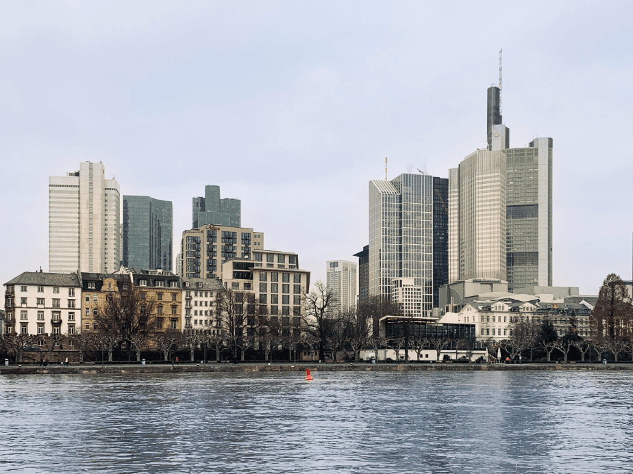 Skyline of Frankfurt with modern skyscrapers and historic buildings by a river, under a cloudy sky.