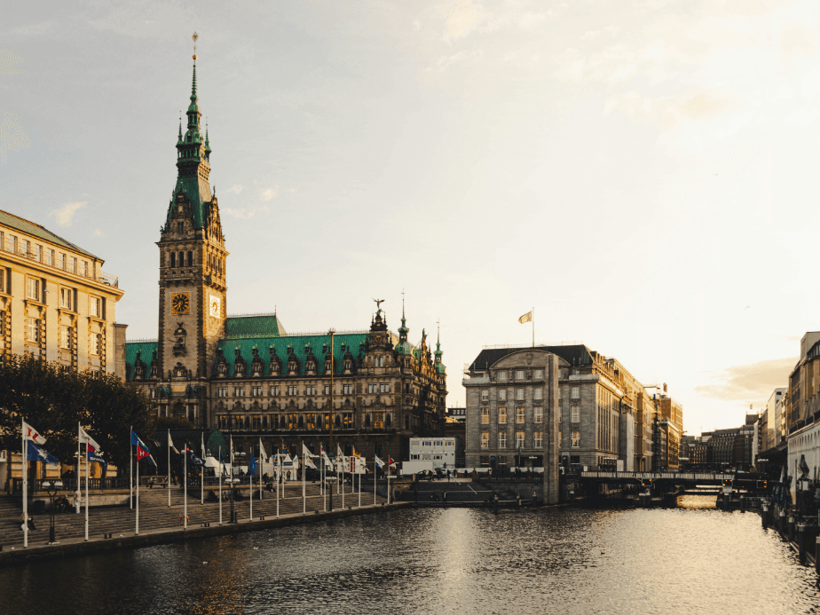 A scenic view of Hamburg City Hall by a canal, with flags lining the water and buildings under a golden sunset.