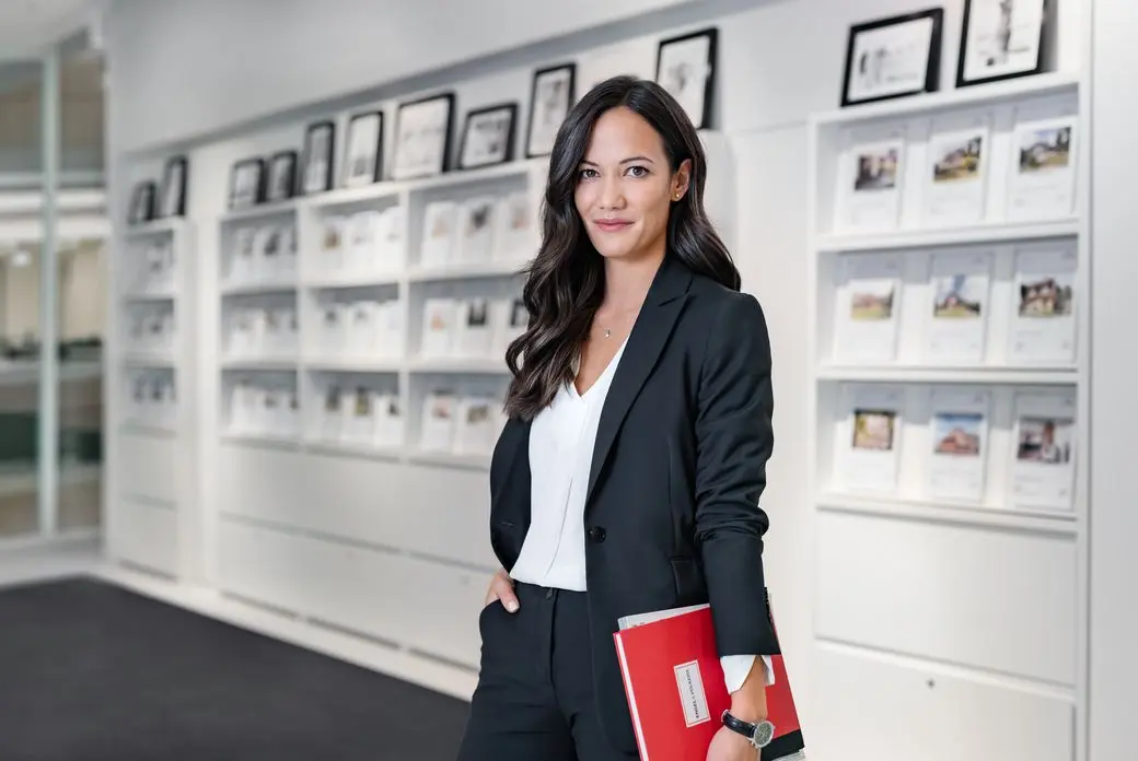 A real estate agent in a black suit holds a red folder in front of a white wall with framed pictures.