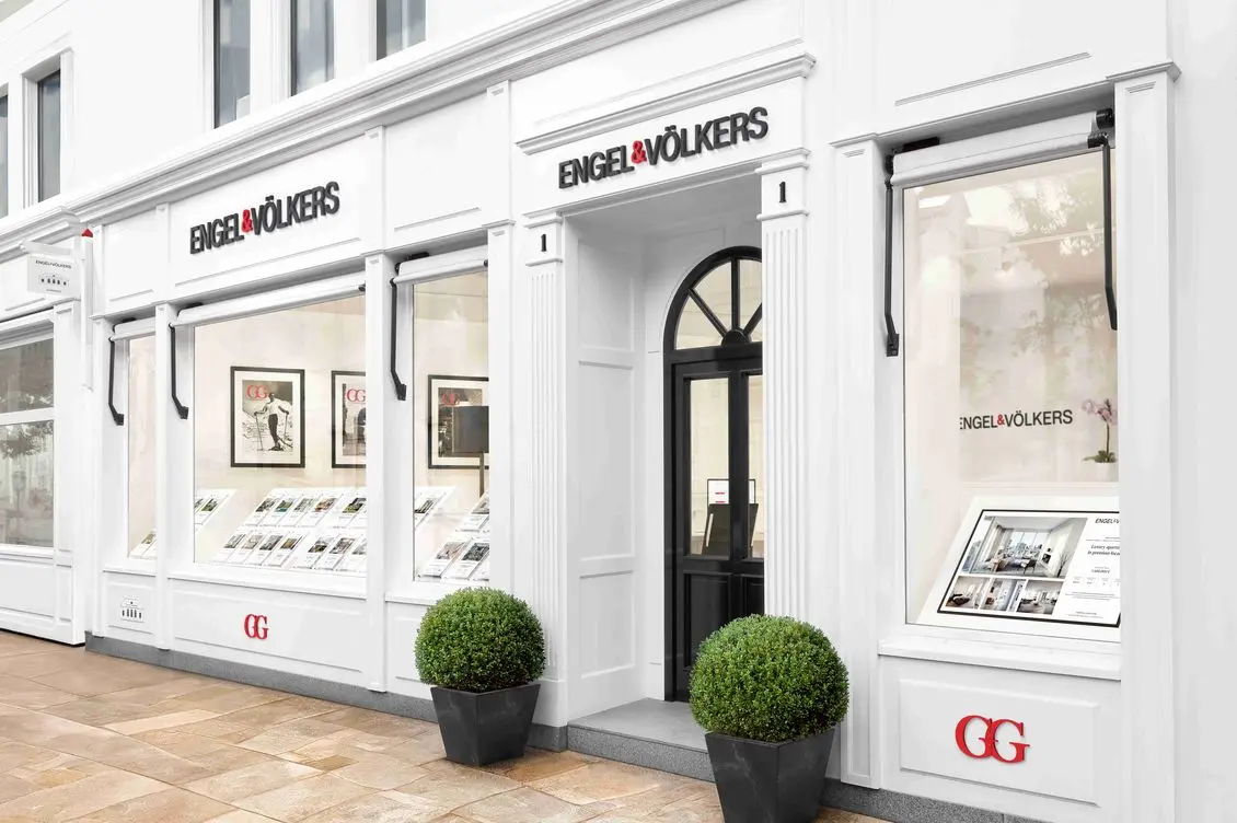 Engel & Völkers κτηματομεσιτικό γραφείο με λευκή πρόσοψη, μαύρη πόρτα και βιτρίνες με καταχωρήσεις ακινήτων. Δύο πράσινοι θάμνοι πλαισιώνουν την είσοδο.