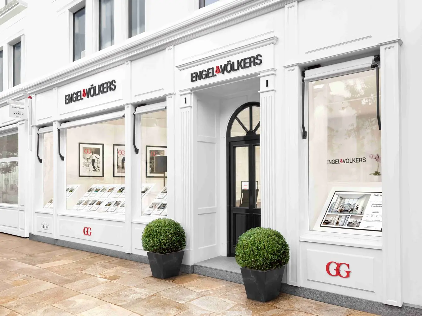 Engel & Völkers real estate office with white facade, black door, and window displays featuring property listings. Two green bushes flank the entrance.