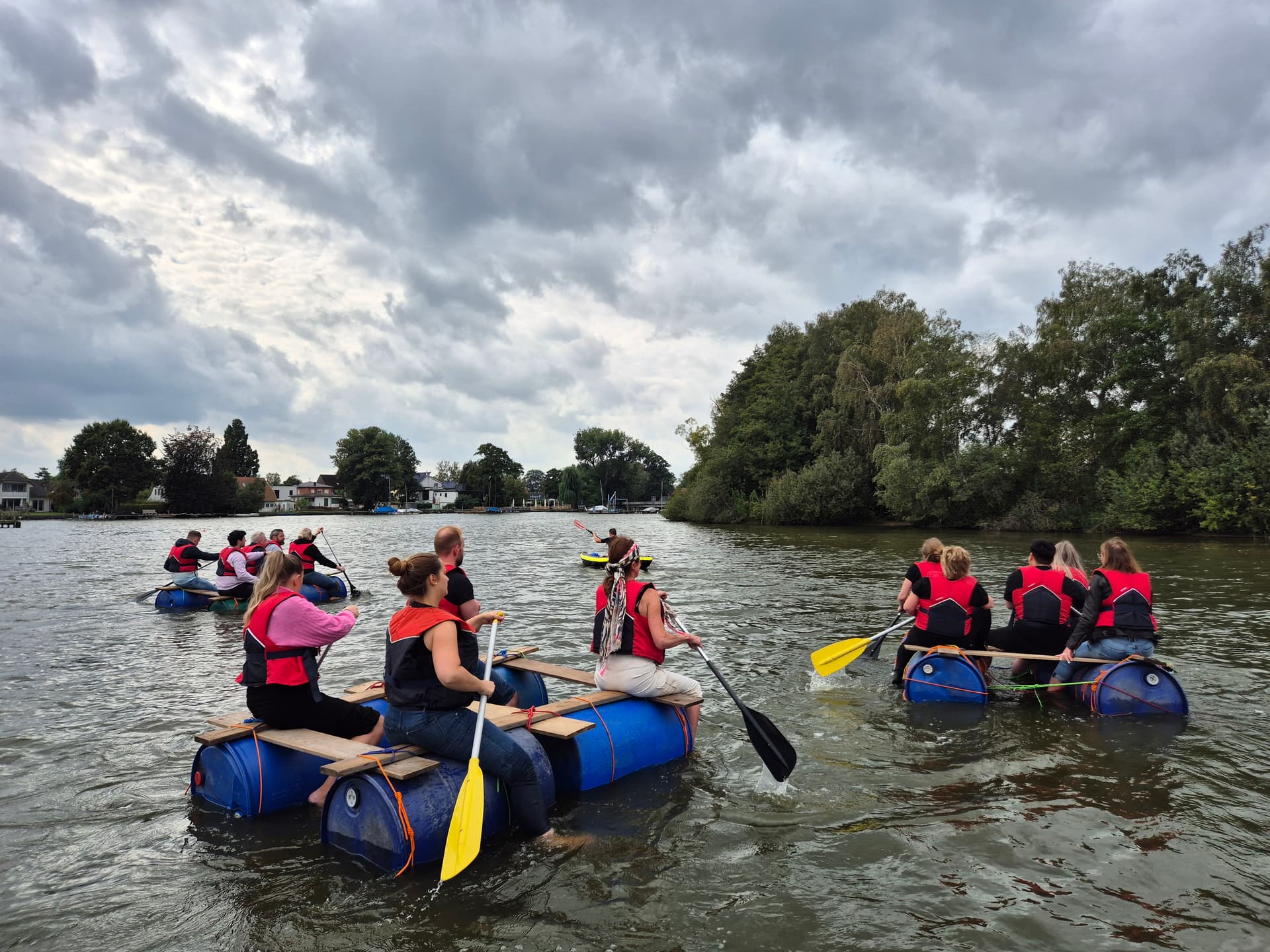 A group of people paddle barrel rafts on a river under a cloudy sky. Trees line the riverbank.