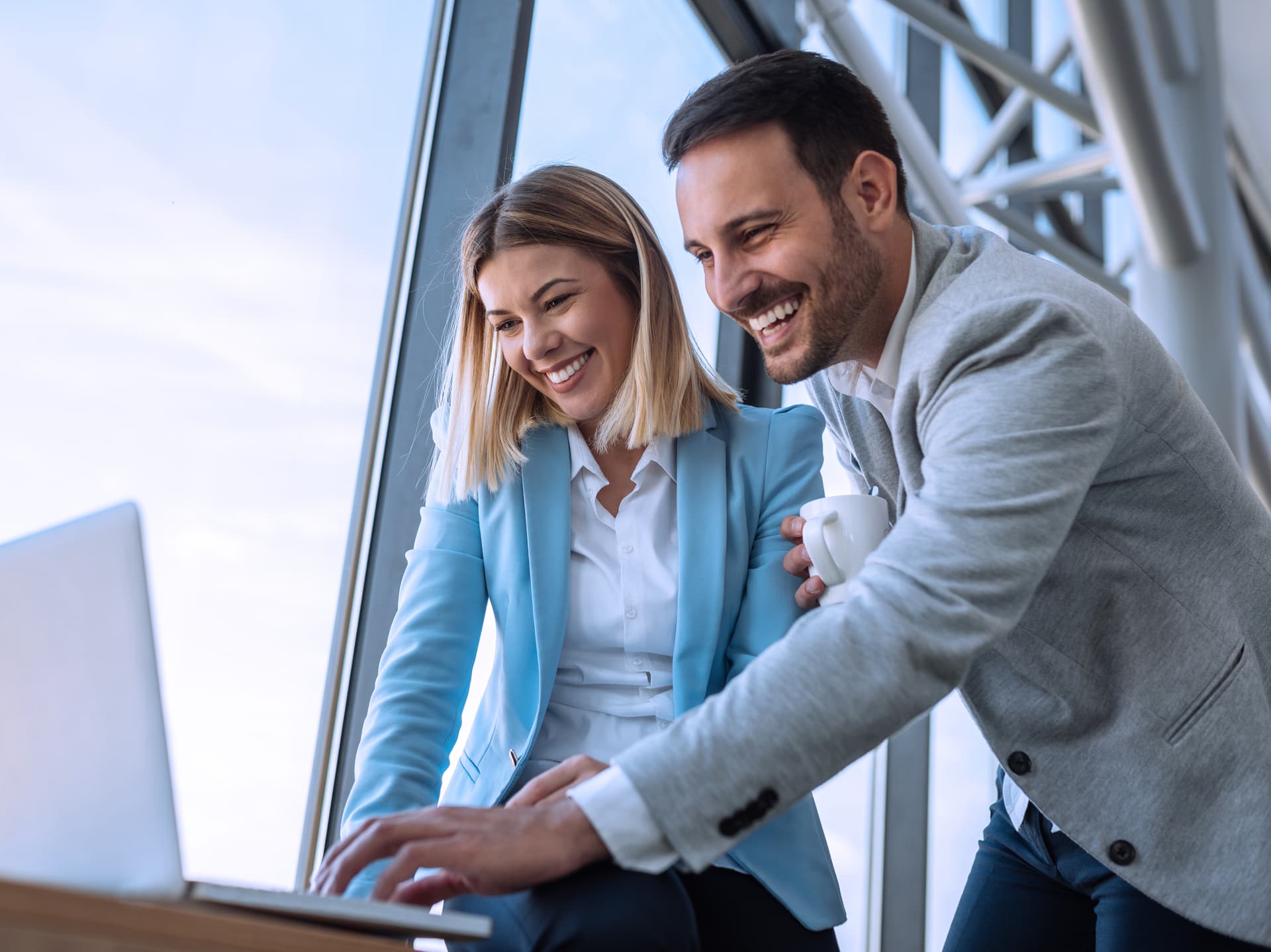 Eine lächelnde Frau und ein Mann schauen auf einen Laptop vor einem großen Fenster. Die Frau trägt einen blauen Blazer und der Mann einen grauen Blazer.