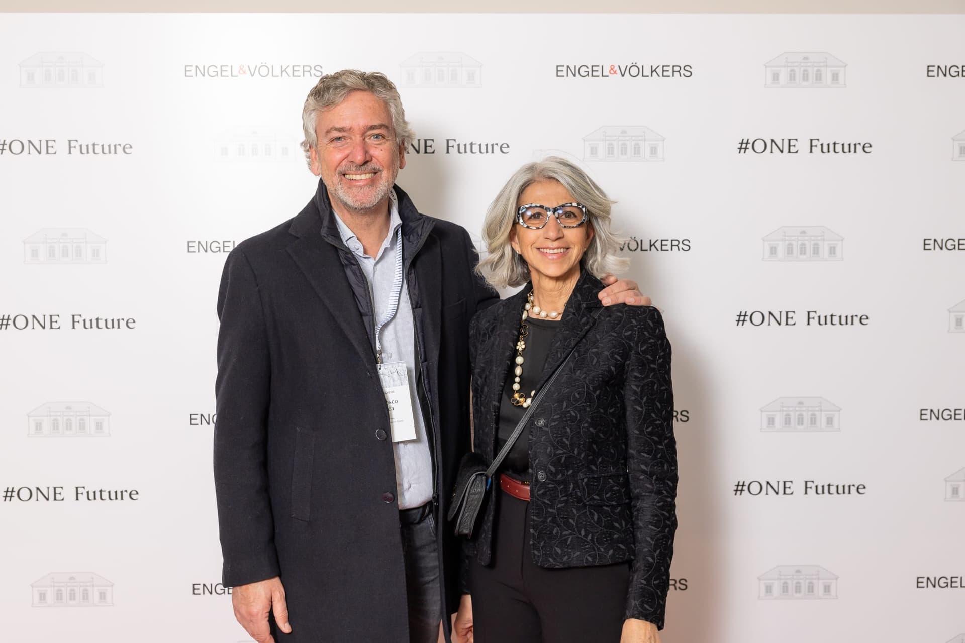 Un uomo e una donna sorridenti posano di fronte a un fondale con il marchio Engel & Völkers. La donna indossa occhiali fantasia e una giacca nera. L'uomo indossa un cappotto scuro.