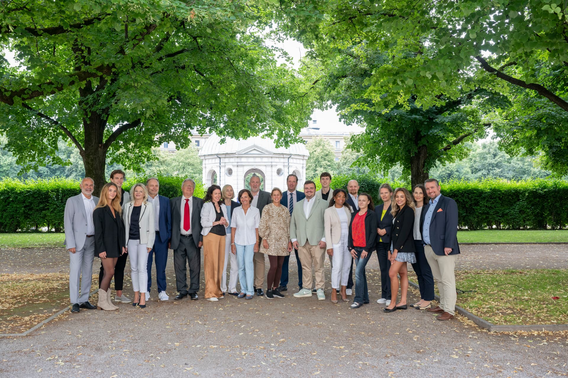 Eine Gruppe von Menschen steht auf einem Pfad unter grünen Bäumen und posiert für ein Foto. Im Hintergrund ist ein weißes Gebäude sichtbar.