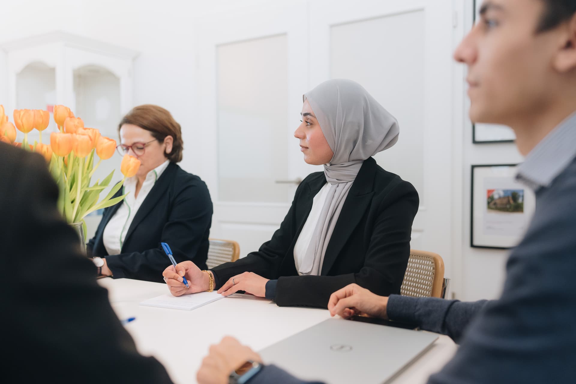 Diverse group at a white table. A woman in a hijab writes with a blue pen. Orange tulips add color to the bright room.