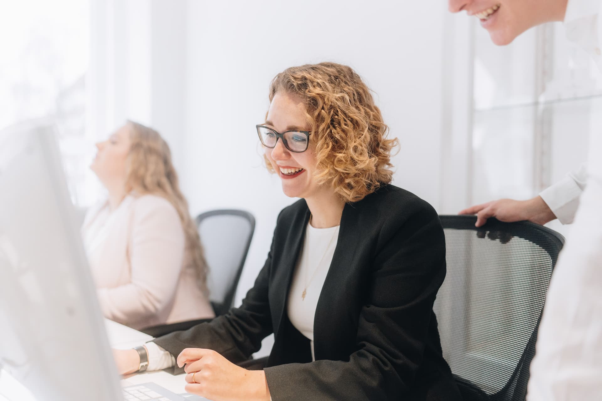 A smiling woman with glasses works at a computer in a bright office. A man stands behind her.